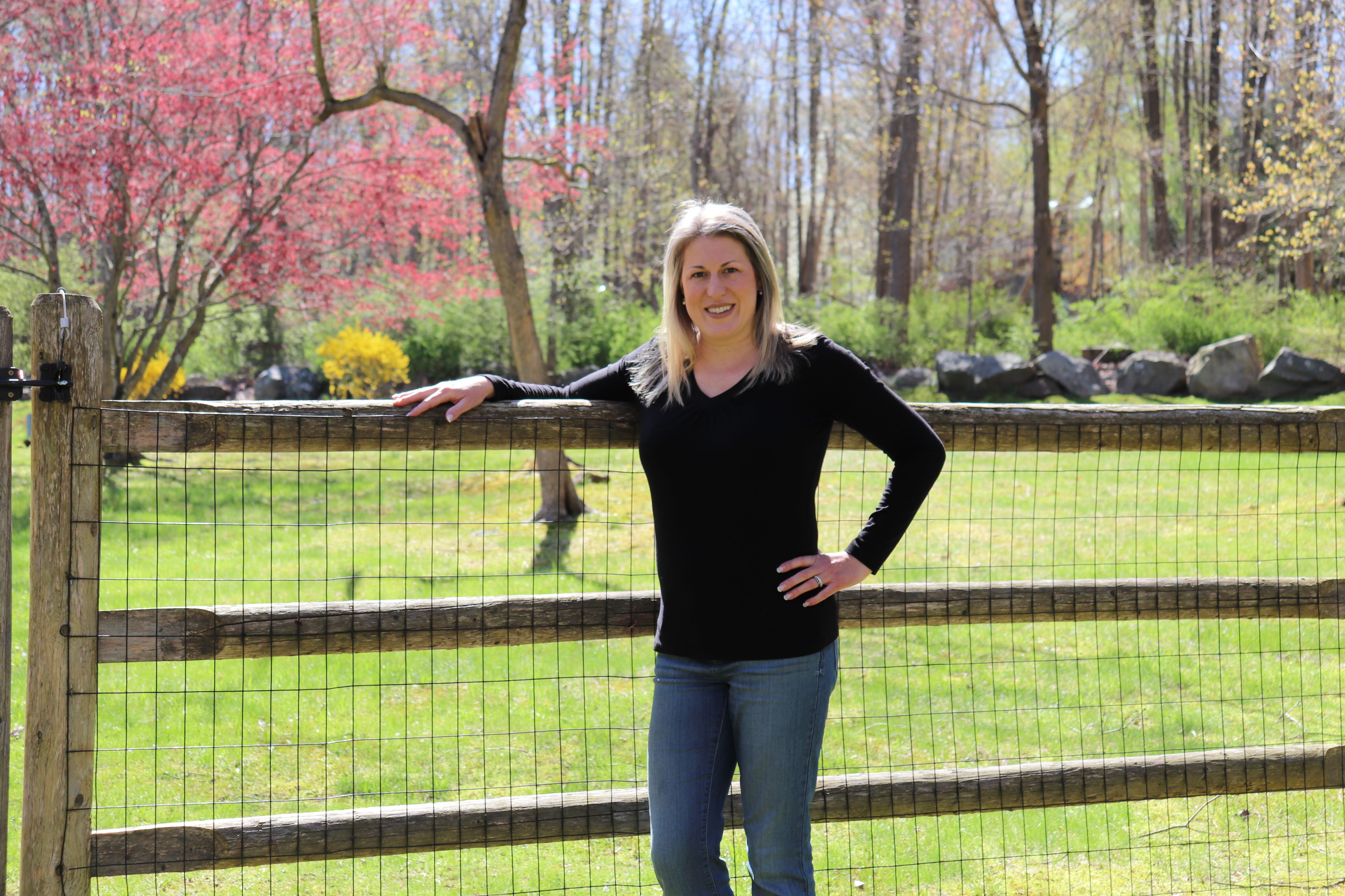 Blond-haired woman in her 40s in a black long sleeve shirt and blue jeans, leaning on a wooden fence.
